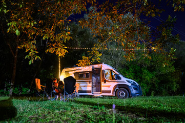 Night camping in nature near river Zeta, Montenegro, with a camper van, chairs, and warm lights under trees, capturing a cozy riverside camping atmosphere and van life experience.
