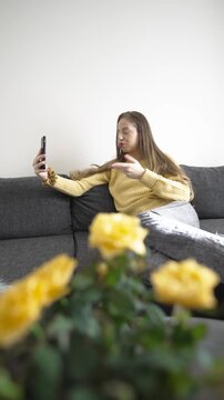 A woman lies on her sofa, making an energetic video call on her smartphone. She gestures animatedly while explaining something, showing a mix of excitement and a slightly disappointed reaction.