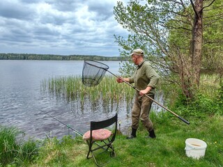 A fisherman uses a landing net to catch fish on the shore of a lake on a summer day.