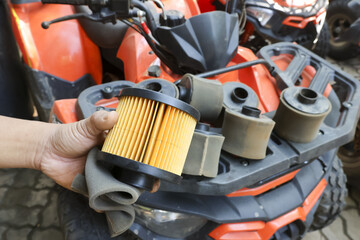 Detail of mechanic hand holding clean yellow air filter for replacement over orange all terrain vehicle with heap of dirty used parts on front rack during seasonal maintenance service © CoreRock