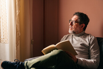 Fototapeta premium Middle aged man contemplating by window with book during quiet moment at home