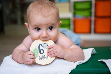 Adorable baby biting six months milestone card while lying on his tummy