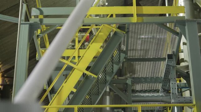 An industrial plant interior with yellow metal structures, large equipment, steel beams, pipes, and mechanisms. A flight of stairs in a large hangar. Production. Working overhead structures.