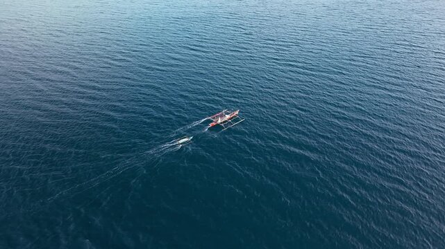Aerial slow motion footage of traditional Filipino bangka outrigger boat towing smaller vessel across deep blue ocean waters. Filmed near Puerto Princesa, Palawan. Shows maritime assistance at sea.