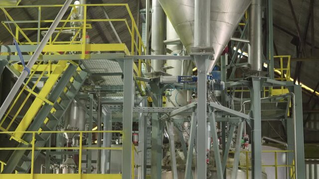 An industrial plant interior with yellow metal structures, large equipment, steel beams, pipes, and mechanisms. A flight of stairs in a large hangar. Production. Working overhead structures.