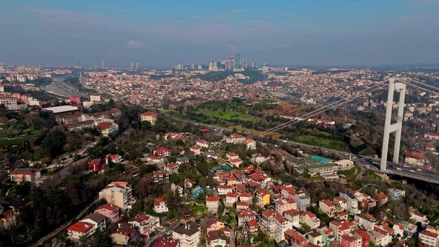Aerial drone footage of the Istanbul Bosphorus coastline, showing dense coastal neighborhoods, red-roof houses, green areas and the bridge, presenting a wide view of the city landscape.