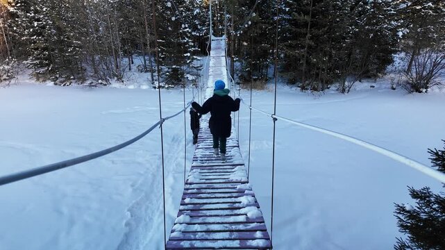 Youngster Walking On Suspension Bridge, Kid In Blue Hat Navigates Wooden Suspension Bridge Over River, Child Wearing Blue Beanie Walks Cautiously On Wooden Suspension Bridge Spanning River