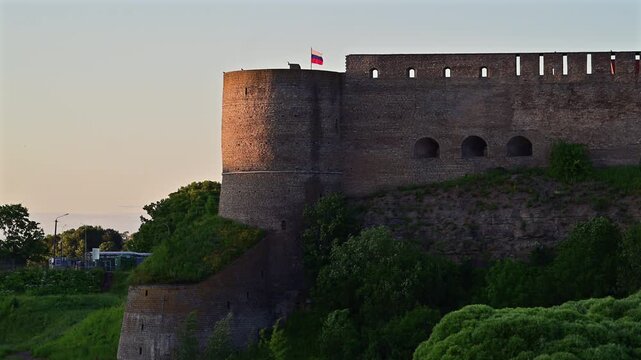 Russian flag atop Ivangorod Fortress on the Russian side of Narva river crossing with Estonia, captured at sunset with medieval stone walls in sharp focus