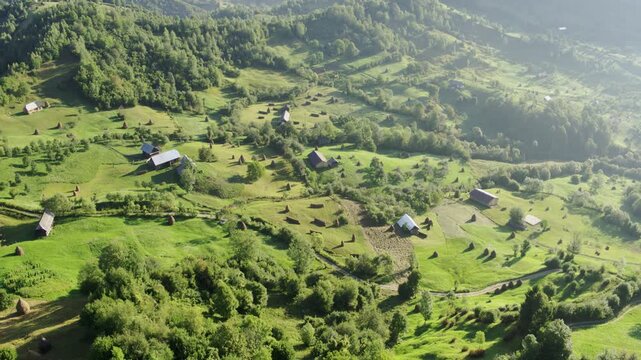 Cinematic drone shot of a traditional pastoral landscape with rustic cottages and haystacks in the Romanian mountains.