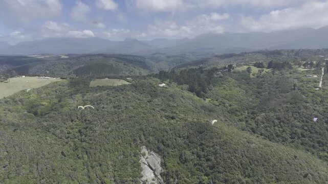 Drone flies backwards from paragliders over forested hll next to coastal highway on a sunny day on the Garden Route in Wilderness, South Africa