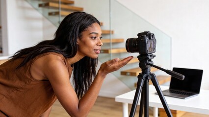 A woman stands near a laptop and tripod, gesturing with her hand. It conveys creativity, content creation, or a behind-the-scenes look at a digital project.