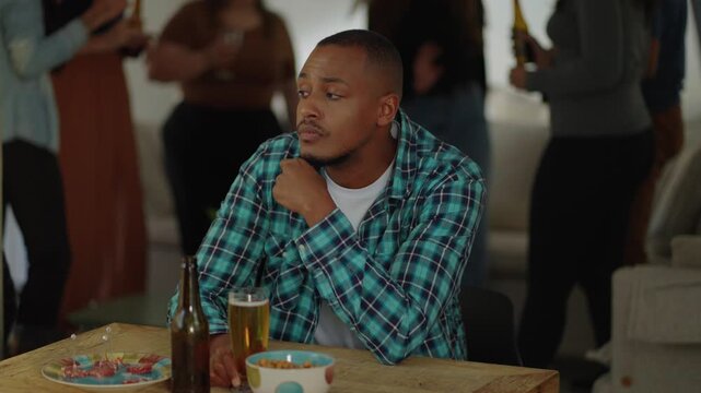Sad upset depressed african black male drinking alone during fun dance party, friends dancing on background. Cultural shock, misunderstanding, language barrier. Apathetic friends
