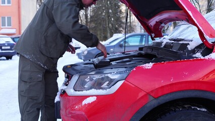 A man checks the engine of a red car in a snowy parking lot. The scene includes several parked vehicles and snow-covered ground.