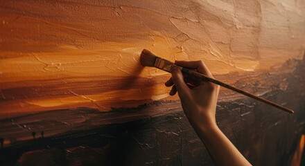A hand holding a paintbrush applying paint to a canvas with warm colors