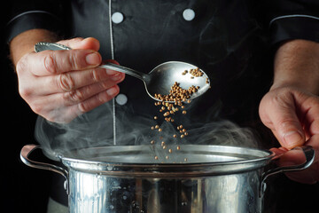 A chef stands in a kitchen, pouring coriander from a spoon into a pot. The steam rises from the pot creating a warm atmosphere. Focus is on the chef's hands and the cooking process