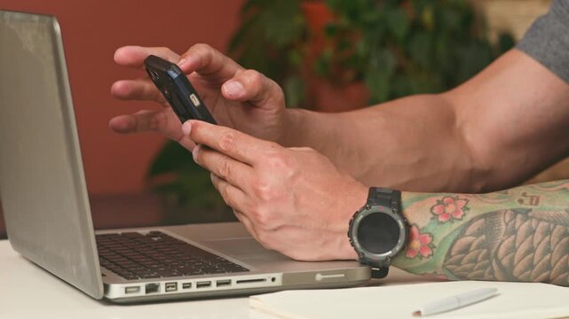 Hands Of A Man With Tattoos Using A Smartphone Next To An Open Laptop