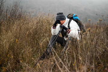 Exhausted and worried hiker in warm clothing sitting alone with backpack and hiking poles in nature on cold foggy winter day. He got lost during hiking.	