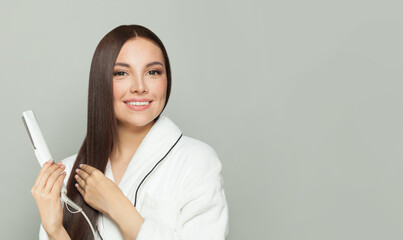 Glorious woman brunette with long straight smooth hairstyle using hair straightener iron on light grey background