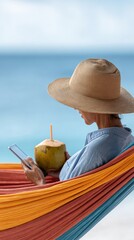Woman wearing a straw hat is relaxing in colorful hammock by the sea, working remotely on her smartphone while enjoying a refreshing coconut drink