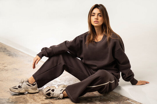 Beautiful, natural woman with long hair posing in studio, wearing comfortable brown tracksuit with an oversized sweatshirt and sporty shoes. Girl is sitting on the floor. Full length photo. 