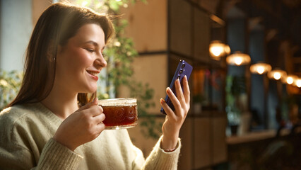 Smiling woman drinking coffee while checking smartphone in warm cafe interior. Concept of coffee shop marketing, lifestyle beverage branding, social media advertising, and remote work.