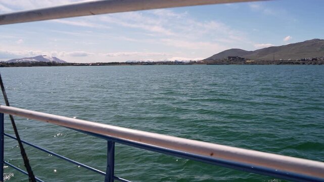 View of lake and mountains from boat on sunny day in nature.Water reflects sunlight as the boat moves gently. Mountains and trees are visible on the horizon under a clear blue sky. People enjoy the ou