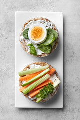 Puffed rice cakes with cream cheese, boiled egg, spinach, carrot and celery on a white plate grey background. Top view