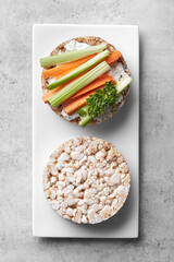 Puffed rice cakes with cream cheese, carrot and celery on a white plate grey background. Top view