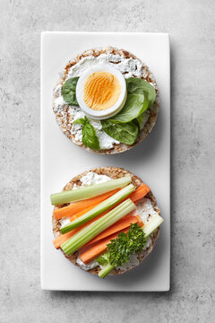 Puffed rice cakes with cream cheese, boiled egg, spinach, carrot and celery on a white plate grey background. Top view