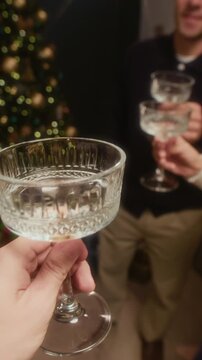 Vertical POV shot of friends clinking champagne glasses in toast for beautiful Christmas holiday celebration at home with glowing and decorated tree standing in living room
