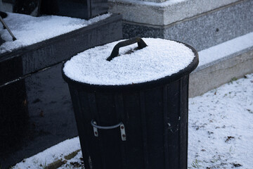 Black cemetery trash bin with a layer of winter snow on the lid among gravestones