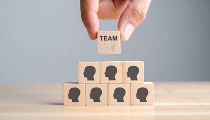 Hand Placing TEAM Block on Wooden Pyramid for Leadership and Collective Success Concept a wooden block with the word "TEAM" atop a pyramid of blocks showing human profile icons