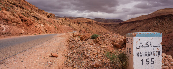 Mountainous road with a distance marker to Marrakech under a cloudy sky. Ouarzazate, Morocco