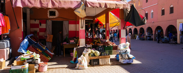 Colorful market stall with produce and goods under red and orange awnings in a sunny plaza. Ouarzazate, Morocco