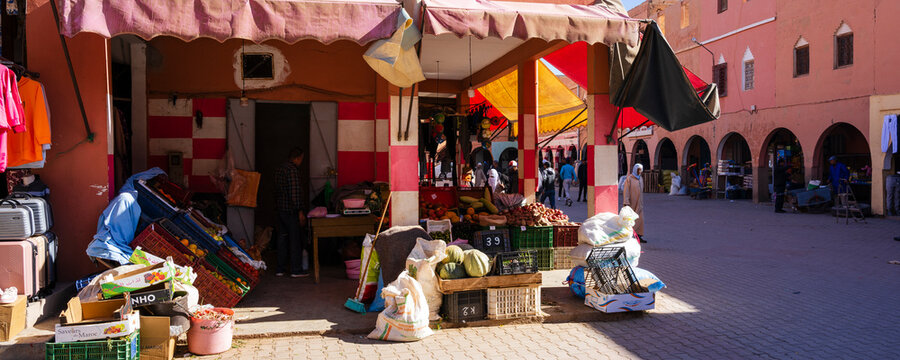 Colorful market stall with produce and goods under red and orange awnings in a sunny plaza. Ouarzazate, Morocco