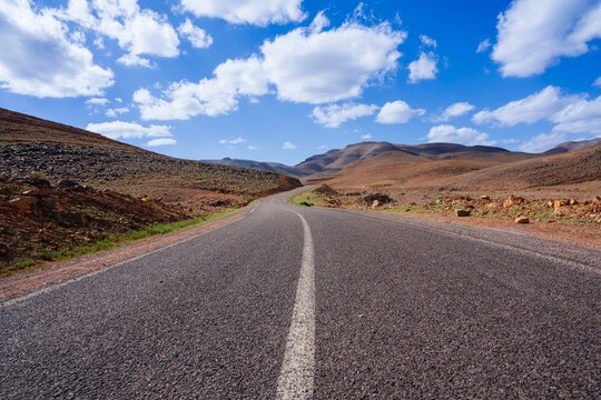 A lonely road stretches through a vast arid landscape beneath a bright blue sky with white clouds. Ouarzazate, Morocco