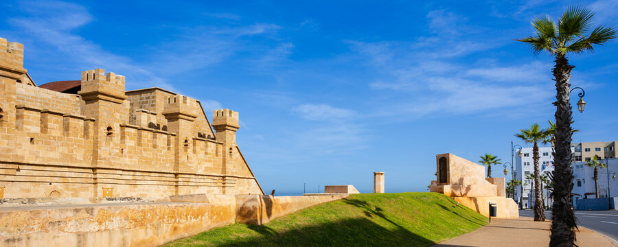 A historic stone fortress stands against a vibrant blue sky, framed by palm trees and grass. Rabat, Morocco