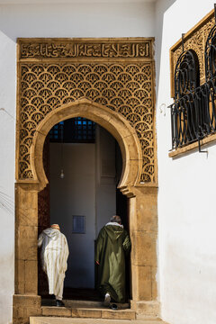Two people in traditional attire enter an ornate arched doorway with intricate carvings. Rabat, Morocco