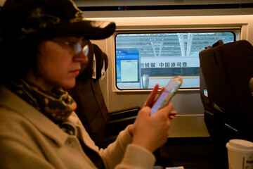 A woman on a train using a smartphone with a view of another train through the window.  Chengdu, Sichuan, China