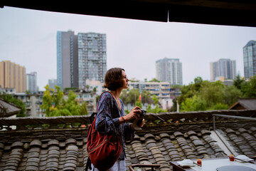 Woman with a camera stands on a rooftop with city skyscrapers in the background. Chengdu, Sichuan, China