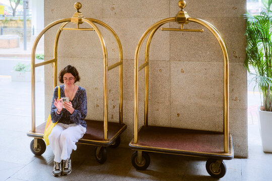 Woman sitting on hotel luggage cart, looking at smartphone, with a second empty cart beside her. Guangzhou, Guangdong, China