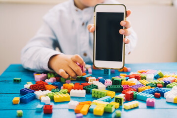 Fototapeta premium Child hand showing mobile phone with blank white screen with colorful toy bricks