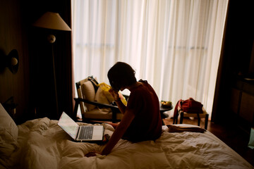 Person sitting on a bed using a laptop in a dimly lit room with sheer curtains. Trip from Guilin to Chengdu, Guangxi, Sichuan, China