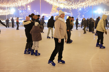 Young girl skating on ice rink under beautiful glowing lights, people enjoying winter activity at public skating arena during evening