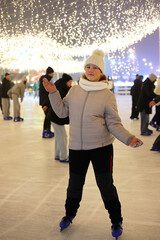 Girl in white hat posing on ice rink with open arms at night, active child having fun during winter skating session under bright lights