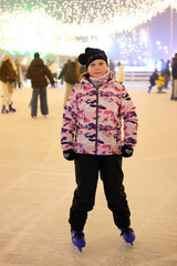 Portrait of young girl in pink jacket standing on ice rink, child ready for skating at evening park under festive garland illumination