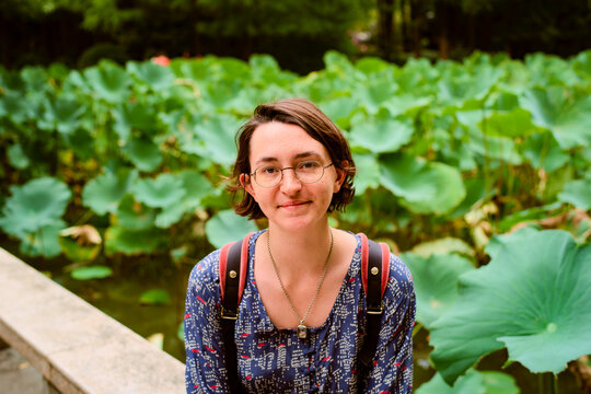 A young adult with glasses and a backpack sits near a pond filled with large green leaves. Shanghai, China