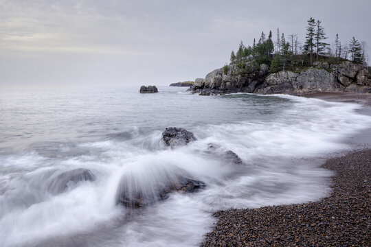 A rocky coastline with waves crashing and trees on the cliff under a cloudy sky. Thunder Bay, Ontario, Canada