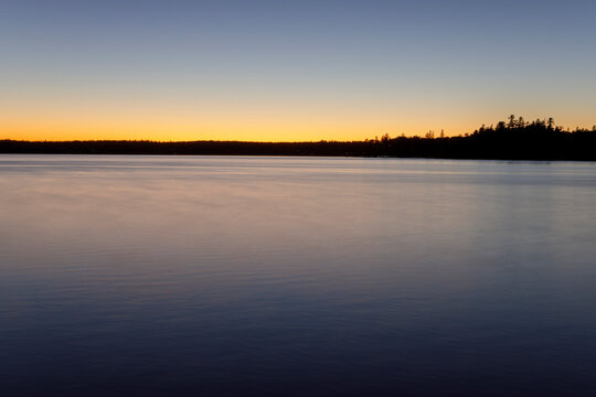 Tranquil sunset over calm lake with silhouette of trees on the horizon. Thunder Bay, Ontario, Canada