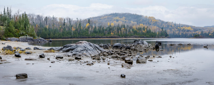 Rocky shoreline by a calm lake with forested hills in the background under a cloudy sky. Thunder Bay, Ontario, Canada